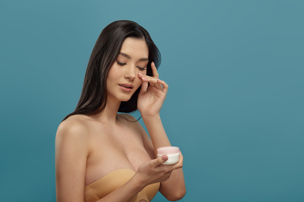 Asian woman holding a skincare product and touching her face